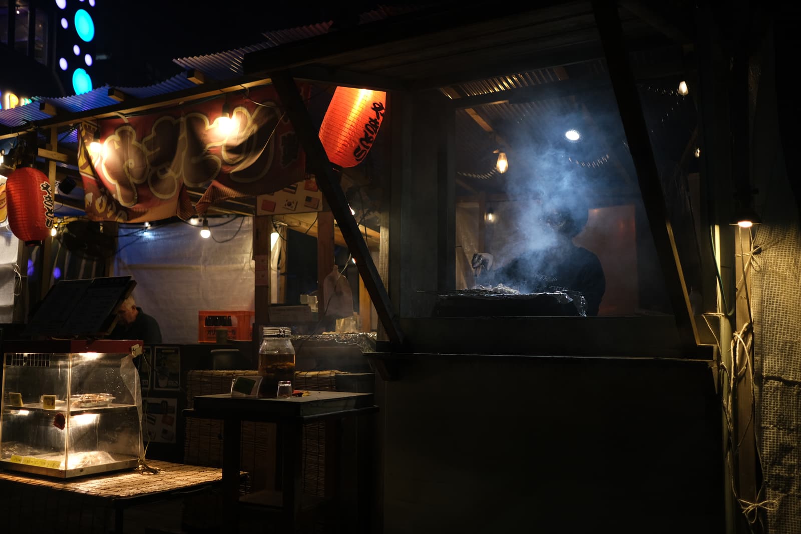 A cook at a food stall at night, smoke rising from the grill